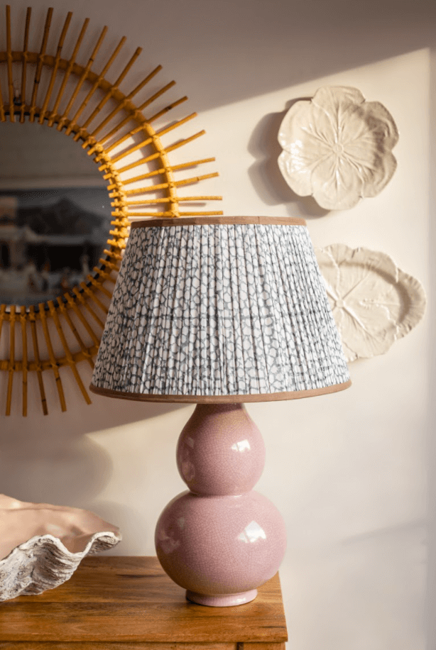 A pink ceramic gourd lamp with an icat lampshade rests on a console table next to a giant clam shell in front of a rattan mirror.