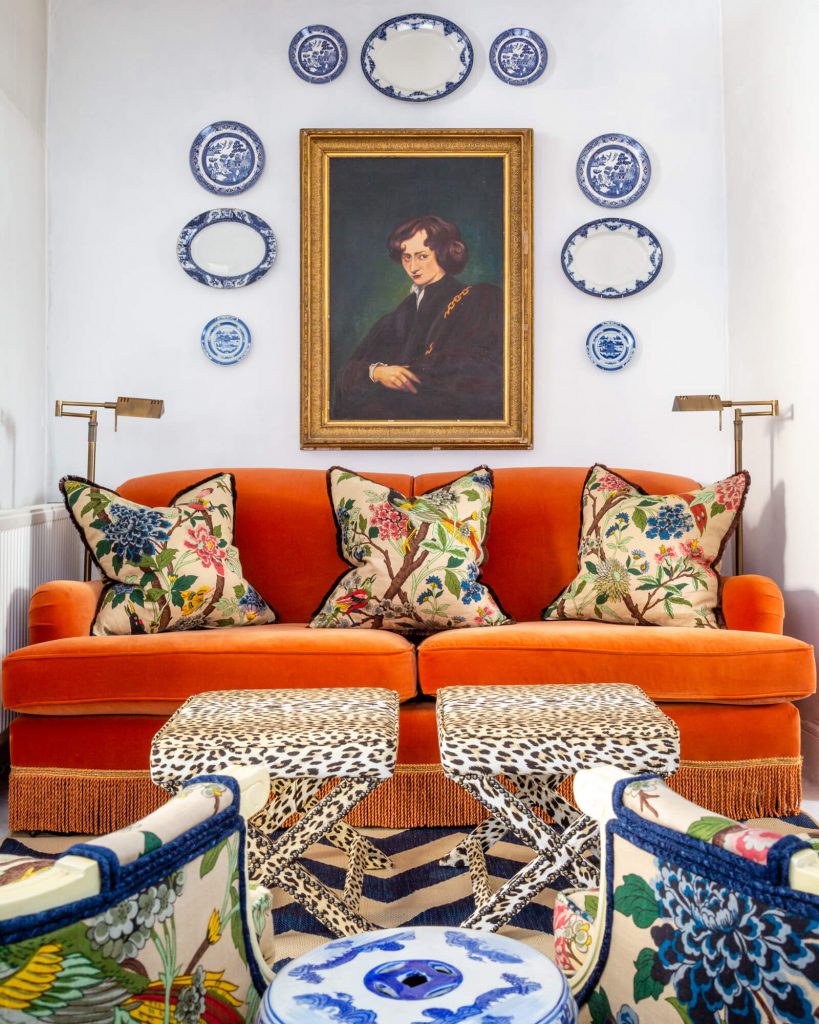 A straight on shot of a vibrant room filled with pattern and print. A burnt orange velvet fringed sofa sits at the centre, with three plush fringed floral cushions sat atop it. Two brass reading lights sit symmetrically on either side of the sofa, and in front are two leopard print upholstered x-frame stools. Above the sofa hangs an oil painting of well dressed courtier and surrounding him symmetrically are blue and white vintage plates. In the foreground sits a large blue and white antique urn and two angled floral armchairs with navy piping. These are in the same fabric as the cushions on the sofa. All of this sits on top of a blue and taupe herringbone jute rug.