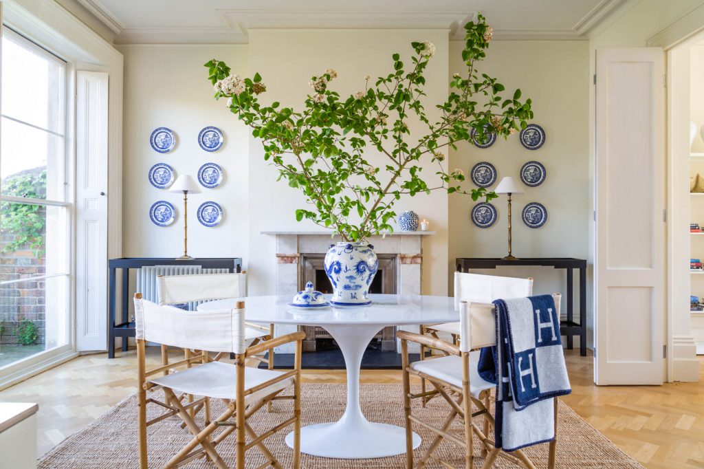 Interior design of a room with a blue and white ginger jar on top of an oval white Saarinen table surrounded by director’s chairs on a sisal rug.